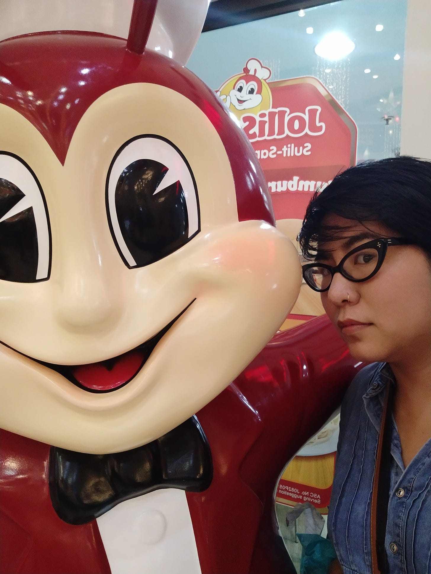    A photo from December 2018. Miranda, with short hair, black cats eye glasses and a nose ring in a denim shirt, poses next to a giant Jollibee statue. His smile is creepily huge. His arm is around Miranda's shoulders.