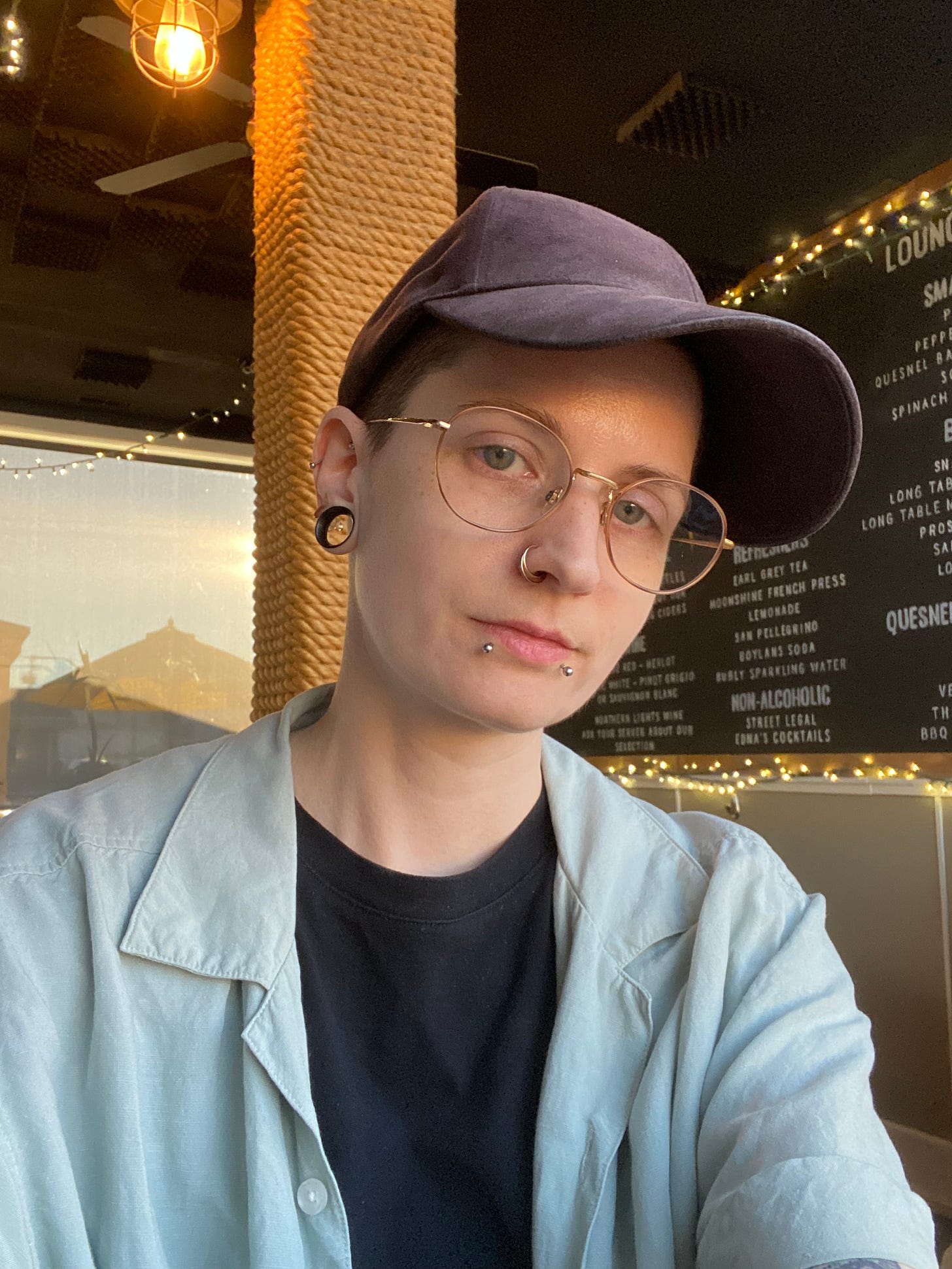 a photo of me, wearing a black crew neck under an open mint green linen button-down and a black baseball cap, in Barkerville Brewery's tasting room. The black menu board is on the wall in the background and there are string lights hung around it.