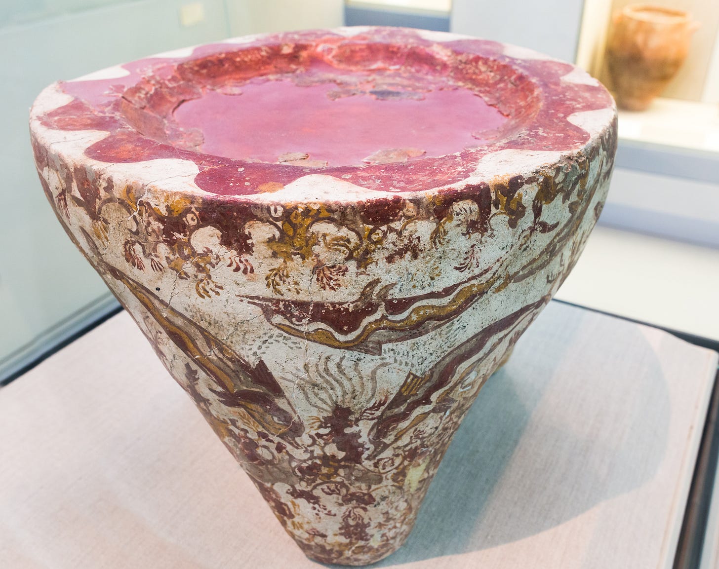 Plastered pumice three-footed offering stand from Akrotiri, ca. 1625 BCE. The stand has a shallow circular depression in the top which is painted red and surrounded by red-painted scalloped decoration. The sides of the stand are painted in a colorful but realistic underwater scene featuring dolphins, coral, and seaweed.