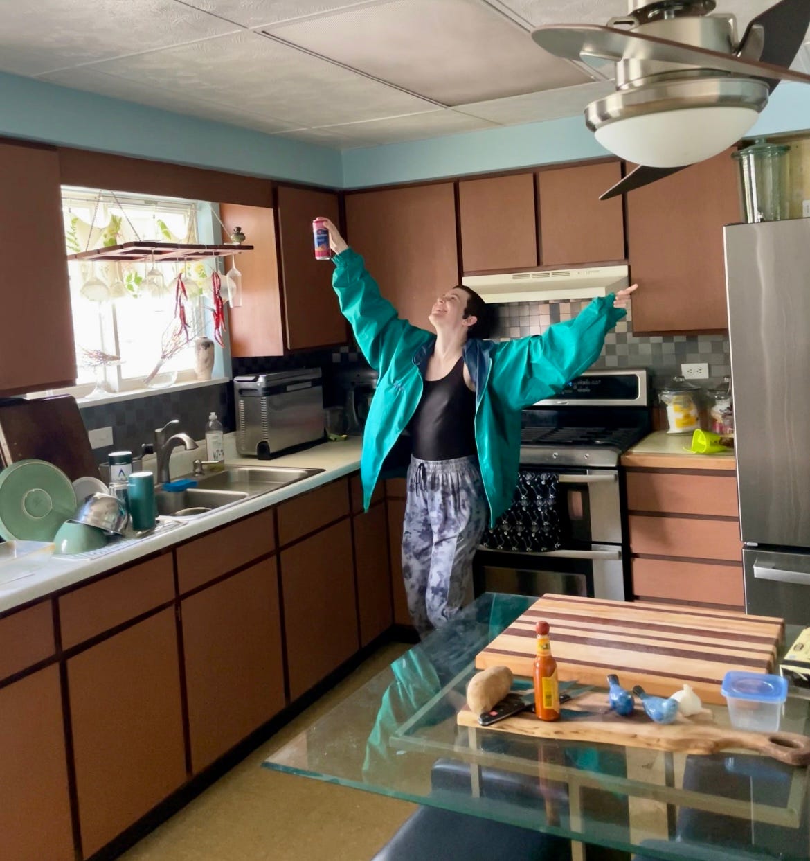 Photograph of Lyss in their kitchen with their arms in the air. They are smiling and dancing while wearing an 80s track jacket and gray sweatpants.