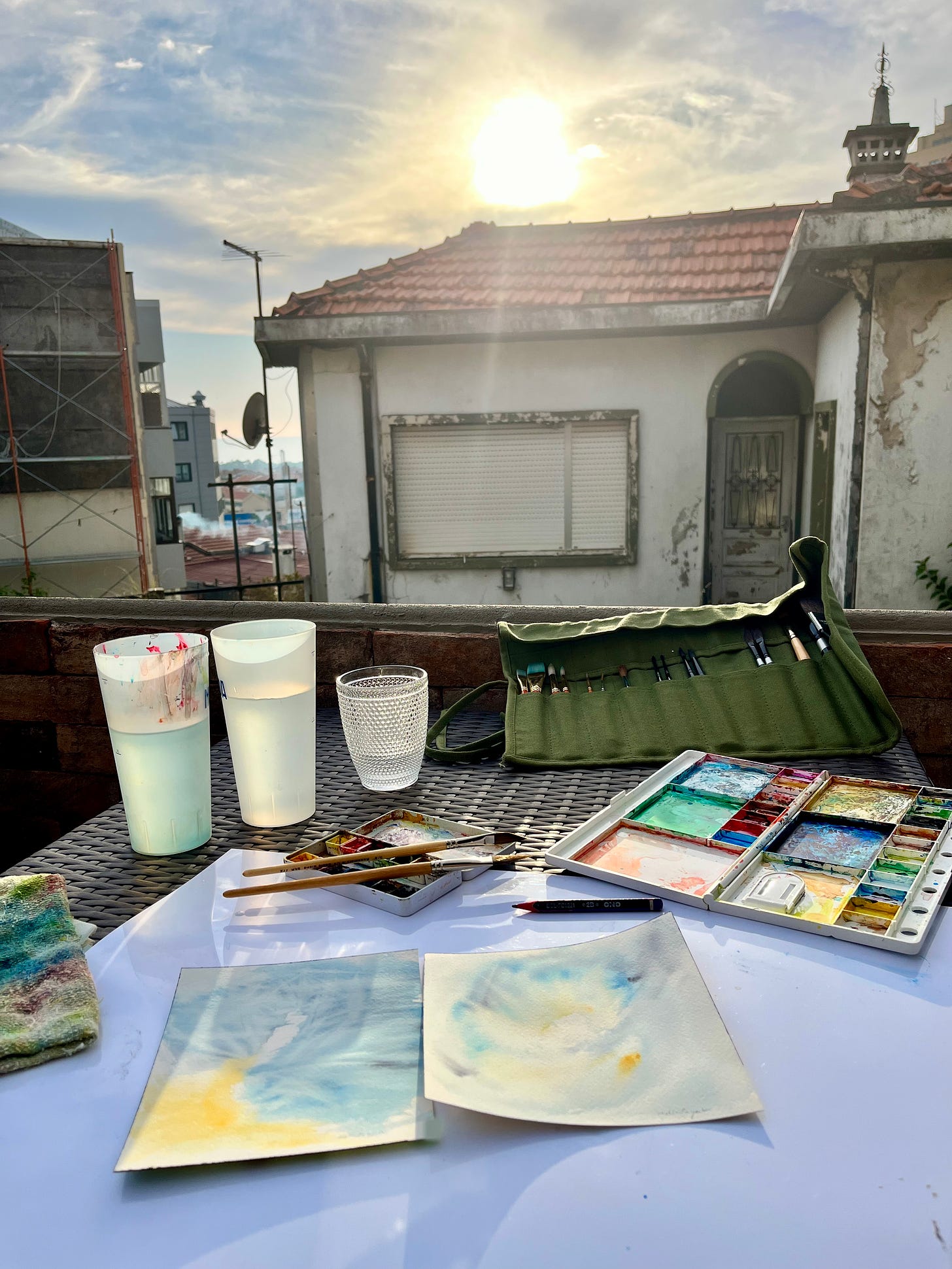 image: an outdoor setting with old houses, evening sky with warm sun, painting with palettes, paints, brushes on the table.