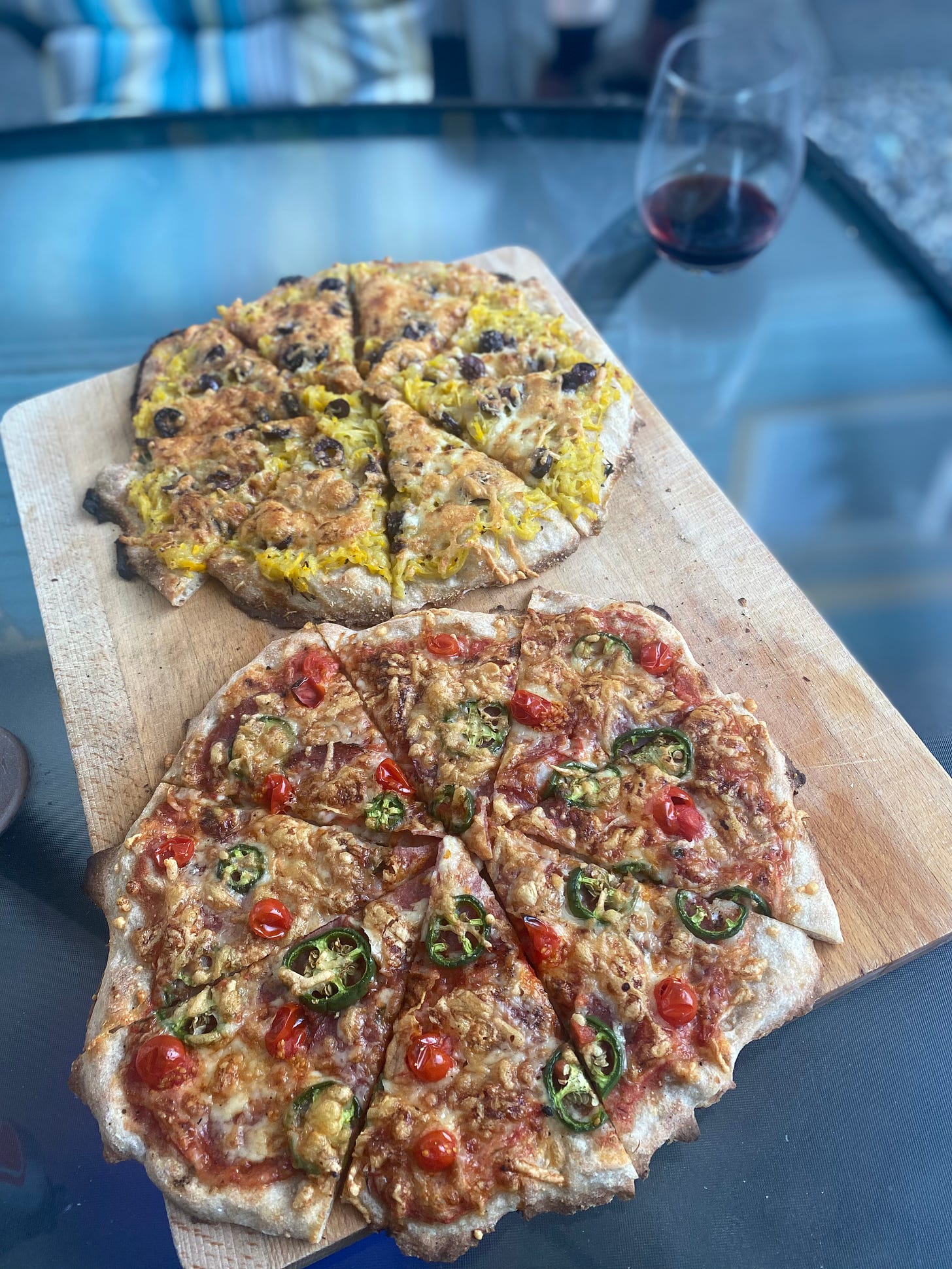 On a wooden cutting board, the two pizzas described above, the salami pizza in the foreground and the zucchini pizza in the background. A stemless glass of red wine is on the table behind them.