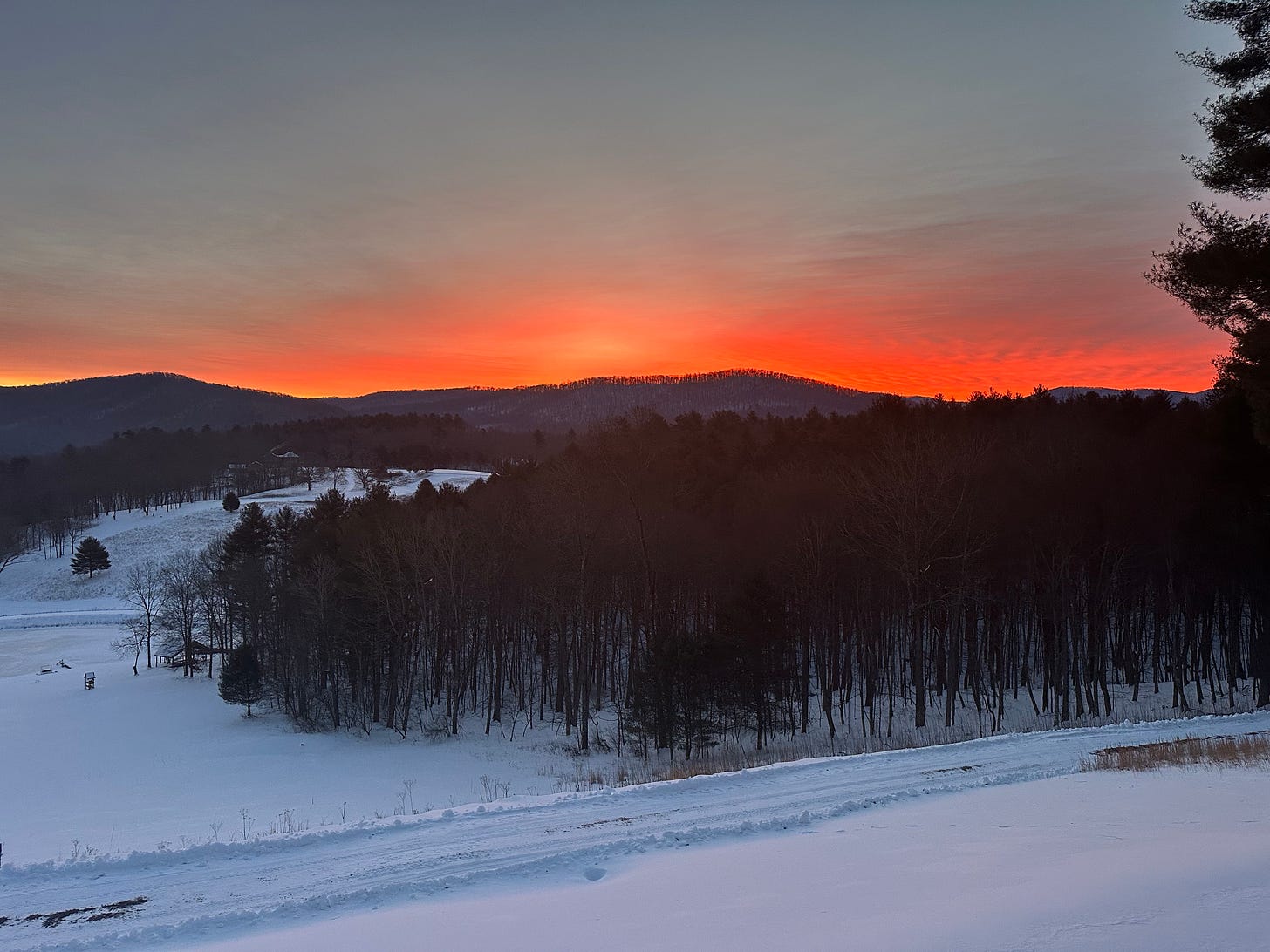 a very bright orange red glow coming just above some dark mountains in the distance, with the snowy fields in front. 