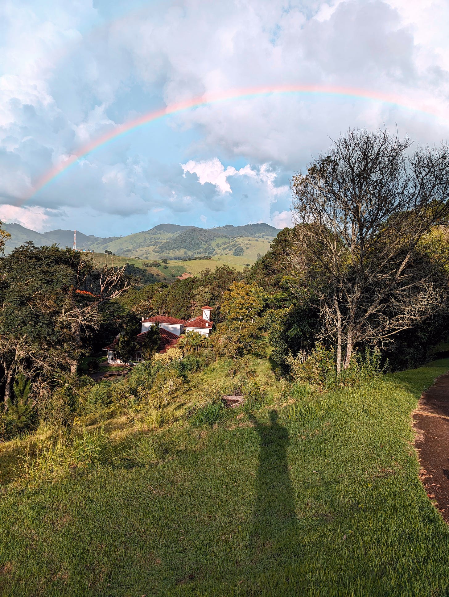 The view of the guesthouse Pousada Dona Marica from the top of a hill, with trees and mountains in the background and a rainbow crossing the sky. The shadow of the photographer can be seen on the grass.