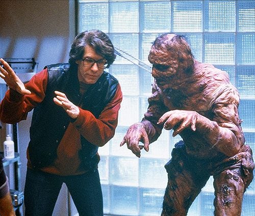 A young David Cronenberg talking with Jeff Goldblum, in full Fly costume, on set of the film. Goldblum is wearing a rubbery, grotesque outfit that looks almost like exposed human flesh that's been melted. Cronenberg is wearing a red sweatshirt and a vest. He has '80s coke bottle glasses.