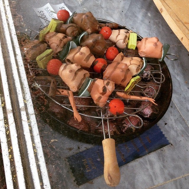 Body parts of Barbie dolls, interspersed with chunks of vegetables on skewer sticks, laying on a grill