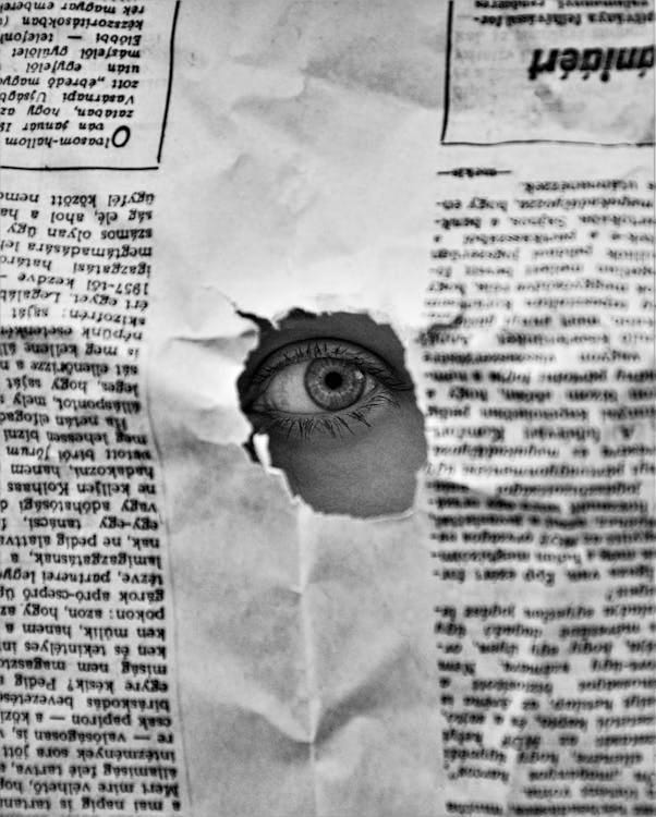 Free A mysterious eye peering through a hole in a black and white newspaper, creating an intriguing visual. Stock Photo