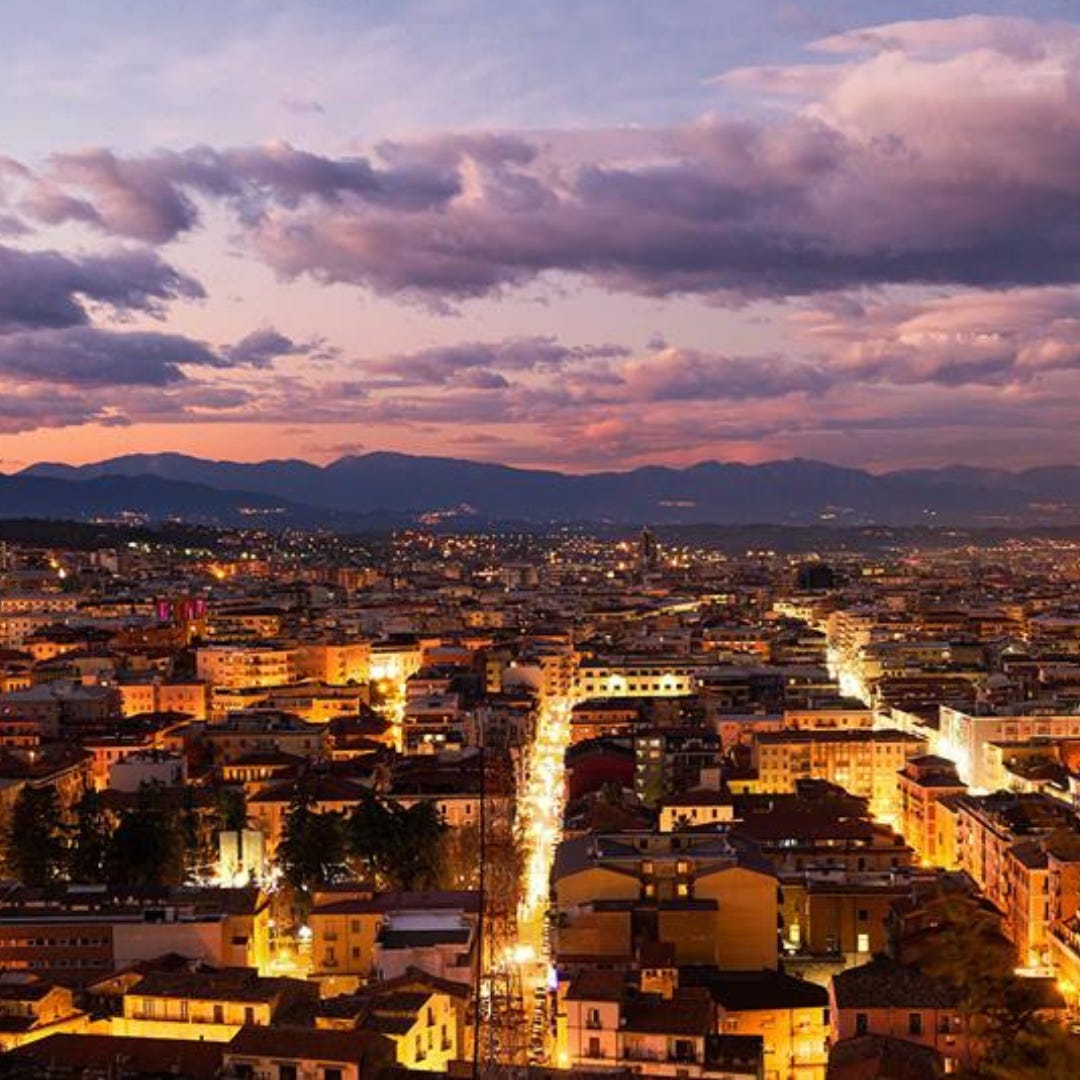 A cityscape at sunset of the old Bruzi capital. The city street lights glow; multi-floor building hug together and pine trees pop out between them. In the distance, a great mountain range rises and falls along the horizon line.