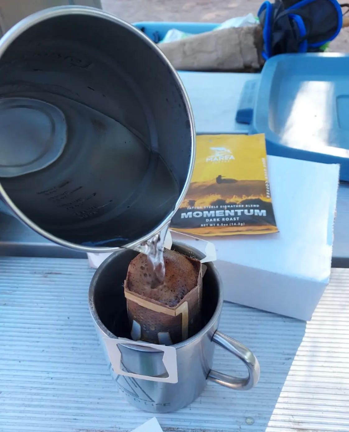 Hot water from a stainless steel Stanley camp pot is being poured over a single-serving pourover coffee in a silver camp mug on a picnic table.
