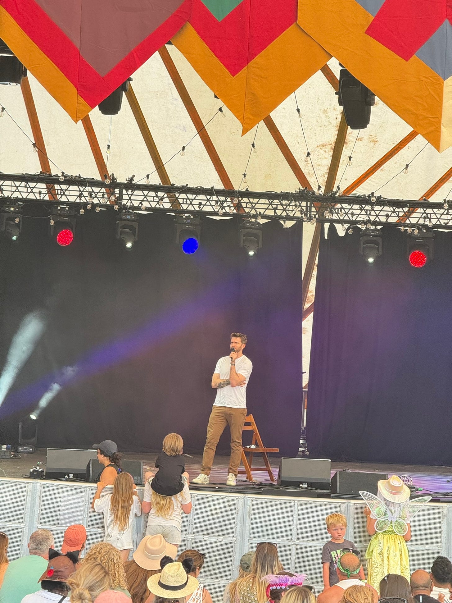 Joel Dommett is standing on stage in front of an audience.  He is wearing a white tshirt and beige trousers plus white trainers.  He is holding a microphone to his mouth and talking.  The stage is in a large purpose built area with gold, pink and grey stripes flags