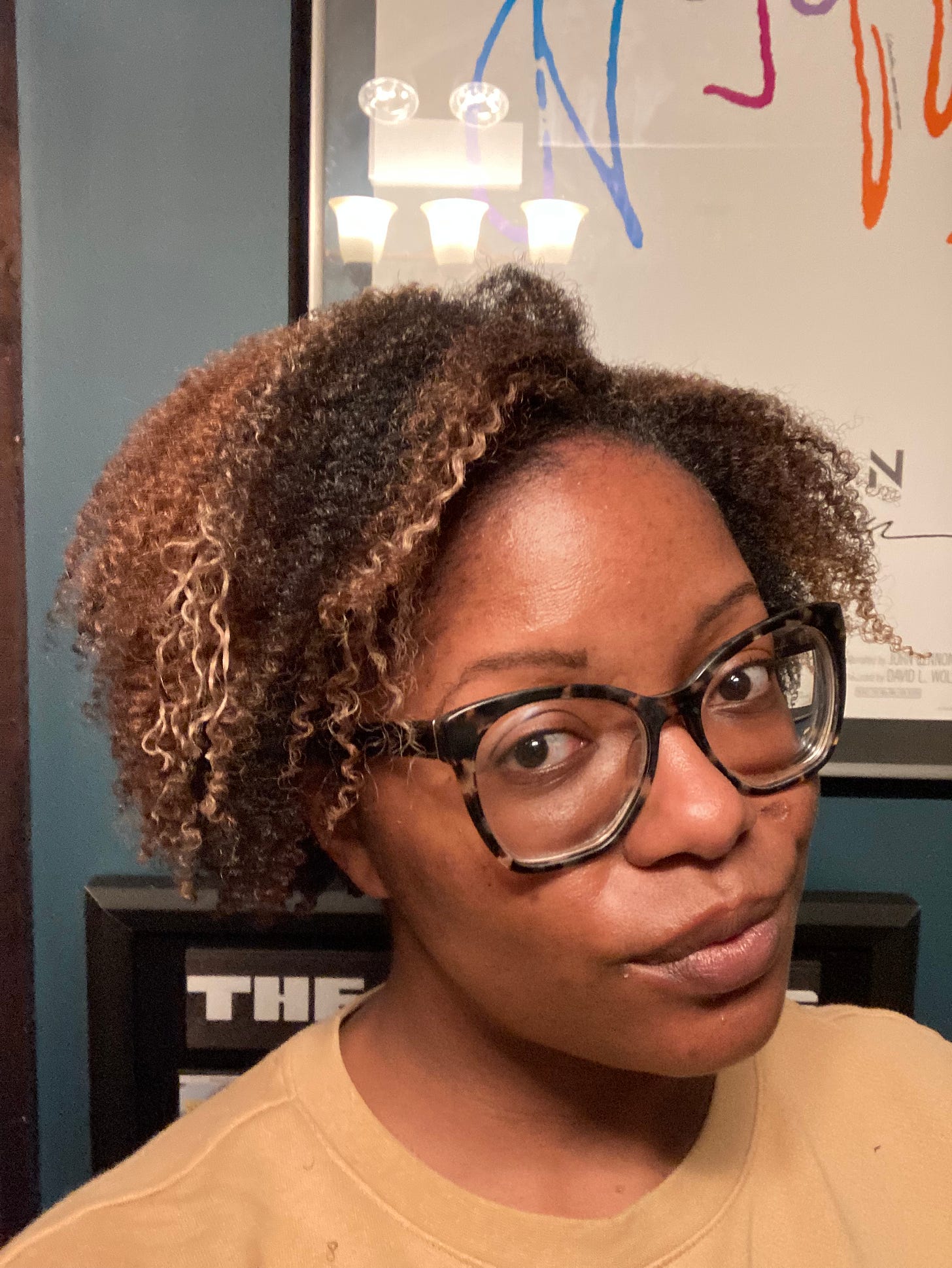 Picture of a Black woman wearing a tan sweatshirt, glasses and a curly haircut with brown and blonde highlights