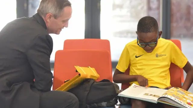 John Todd, head of Charterhouse Nigeria, in a grey suit leans forward in a seat towards a schoolboy in glasses, wearing a yellow Charterhouse Lagos sports shirt, reading a science book