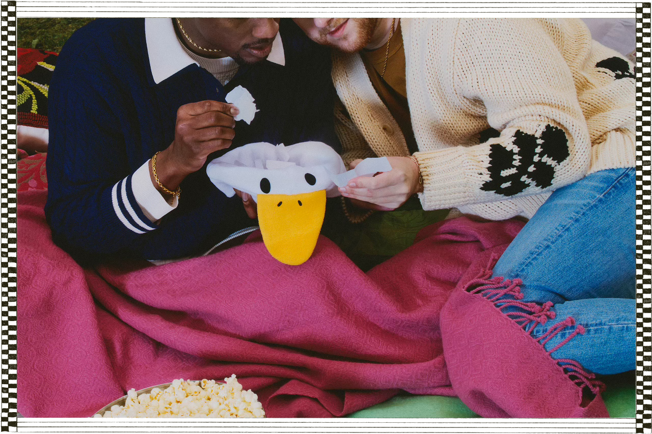 Sam and Tony sit closely under a pink blanket, pulling slips of paper from a plush duck-shaped hat as they watch a film outdoors. A bowl of popcorn rests in front of them.