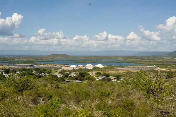 A cluster of white buildings in the distance beyond which lies a blue bay and distant hills.