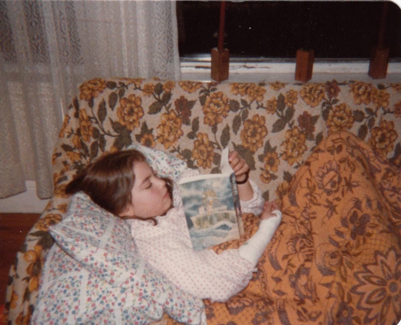 a child with white skin and brown hair is on a floral printed couch reading a book. her right arm is in a cast