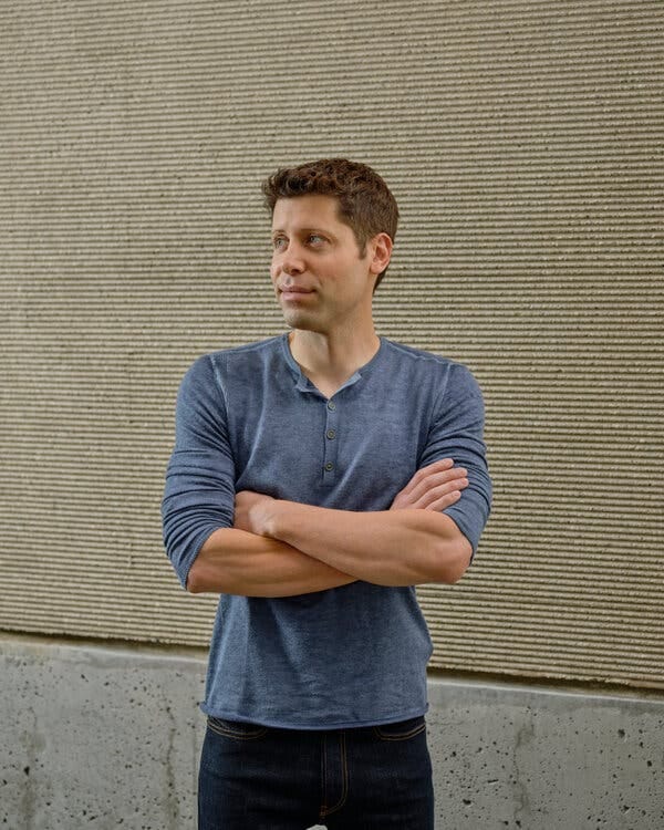 Sam Altman crosses his arms while standing in front of a blank wall. The sleeves of his blue Henley shirt are pushed up above his elbows.
