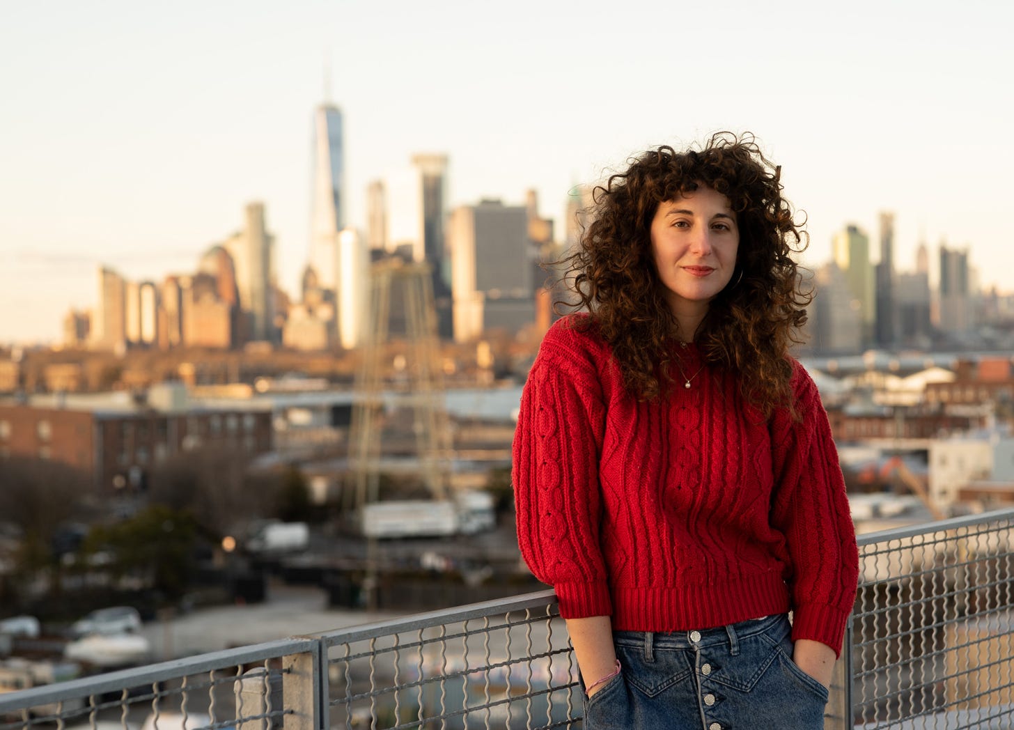 Writer Amanda Chicago Lewis stands on a balcony with the New York City skyline in the background. She's wearing a very cozy-looking red sweater.