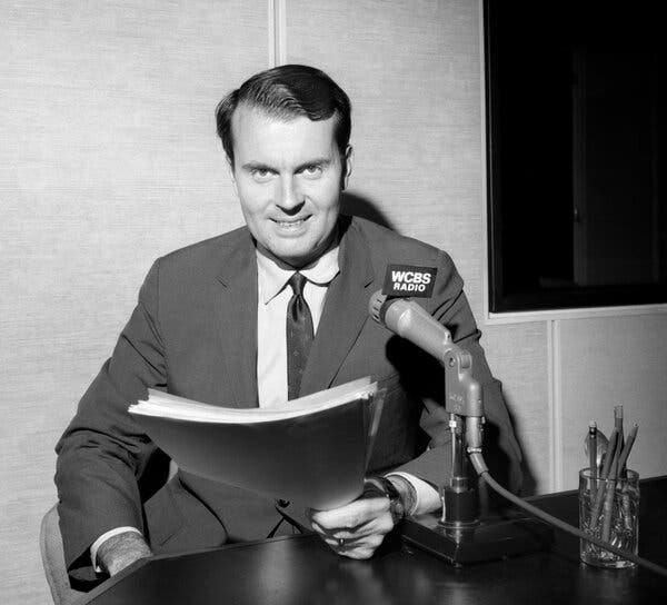 A black-and-white photo of a young Charles Osgood in a studio. seated at a microphone that says “WCBS Radio” and holding a script. His hair is slicked back and he is wearing a jacket and tie and smiling.