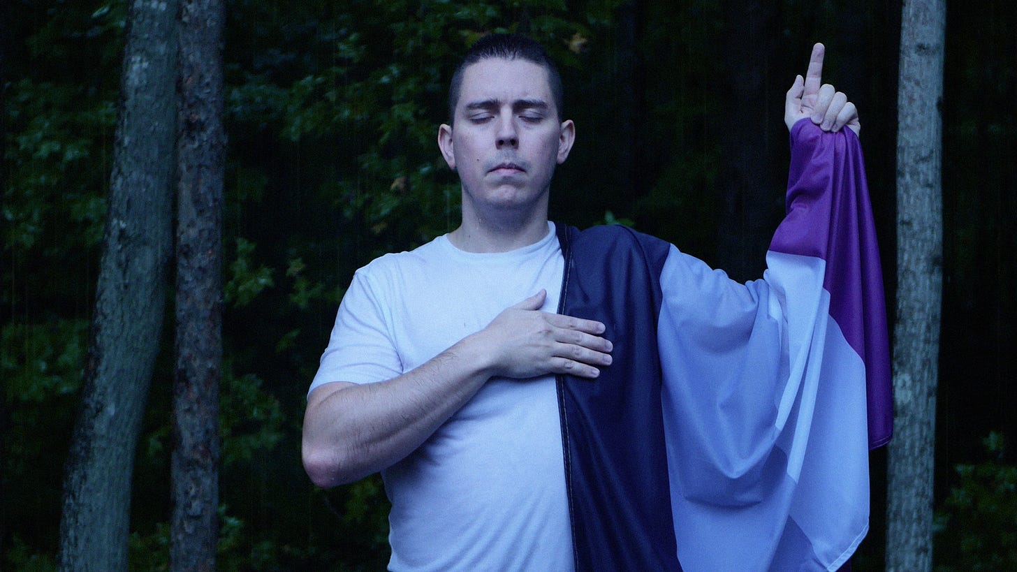 The author stands outside with a dark forest in the background. He's wearing a white short-sleeve shirt with the asexual flag draped over his left arm. His left hand points to the sky, and his right arm folds over his chest with the hand thereof resting on his heart. He wears a contemplative expression with his eyes closed.