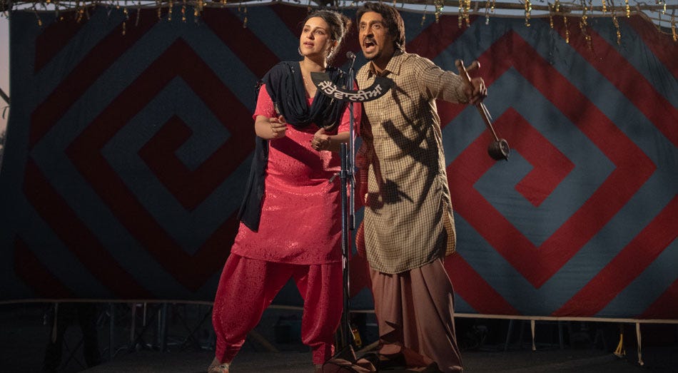 A still from Hindi film, 'Amar Singh Chamkila'. Amarjot (Parineeti Chopra) in a pink salwar-kurta and black duppatta, and Chamkila (Diljit Dosanjh) in beige kurta-tehmat (lungi) and holding a thumbi (musical instrument) singing into a standing mic in what looks like a stage.