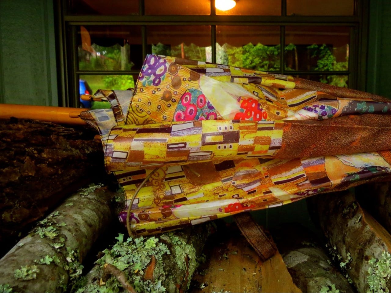 a color photo of an umbrella with a busy, colorful design, laying on top of a stack of wood that has a lot of green lichen growing on it. Behind is a window and an inner area with an overhead light visible. 