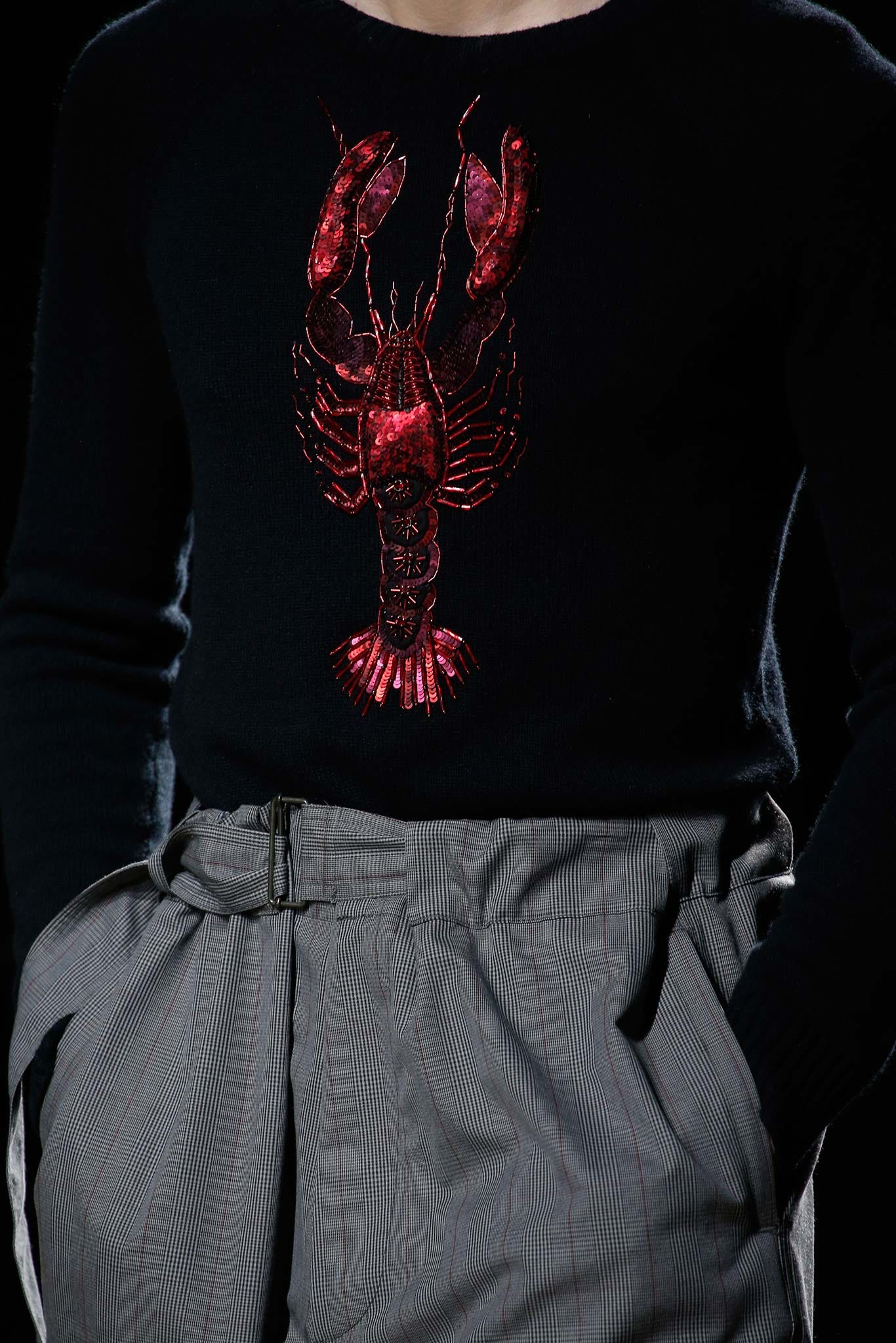 Close-up detail of a navy sweater, with a large embroidered red lobster made of sequins that fills the chest. Close-up detail of a navy sweater, with a large embroidered red lobster made of sequins that fills the chest.