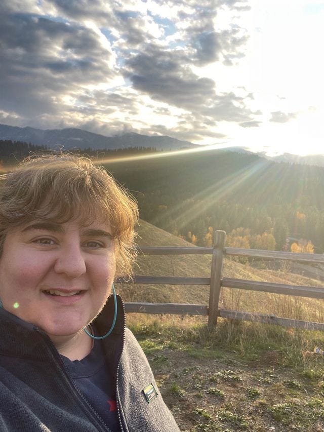 Selfie of Amy, with messy hair, wearing a navy fleece jacket, in front of a wooden rail fence, with a setting sun and mountain valley in the background.
