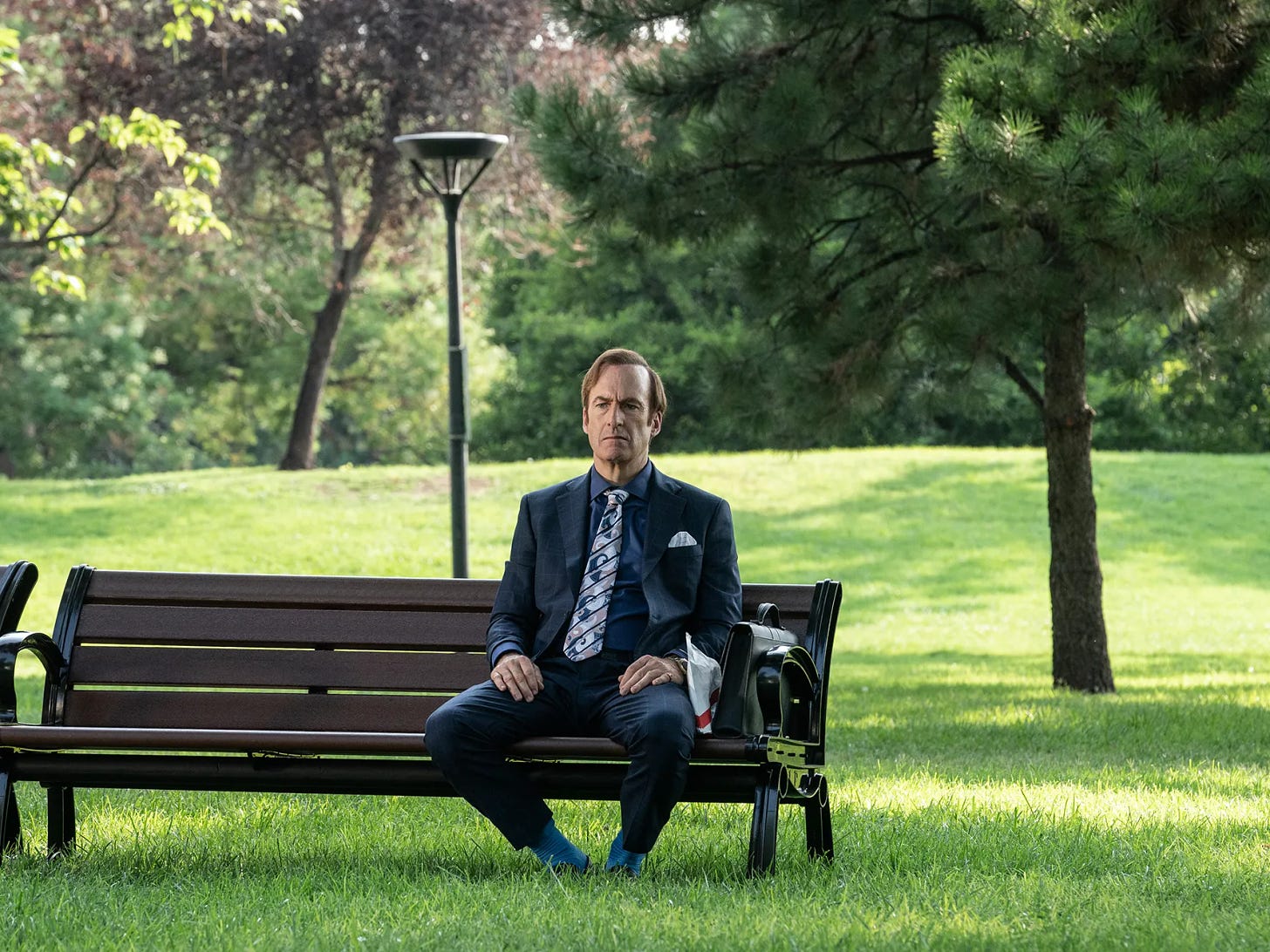 Bob Odenkirk as Jimmy McGill in a suit and tie sitting on a park bench in the middle of a lush green park in the final season of Better Call Saul