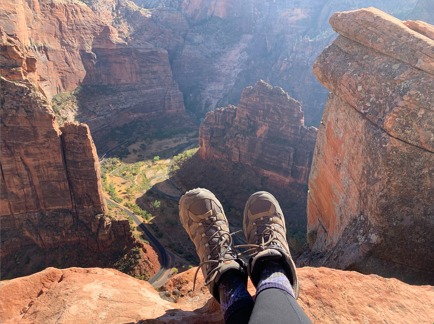 Crossed feet in a pair of hiking boots rest against a cliff overlooking a huge drop over a cliff and a verdant valley below.