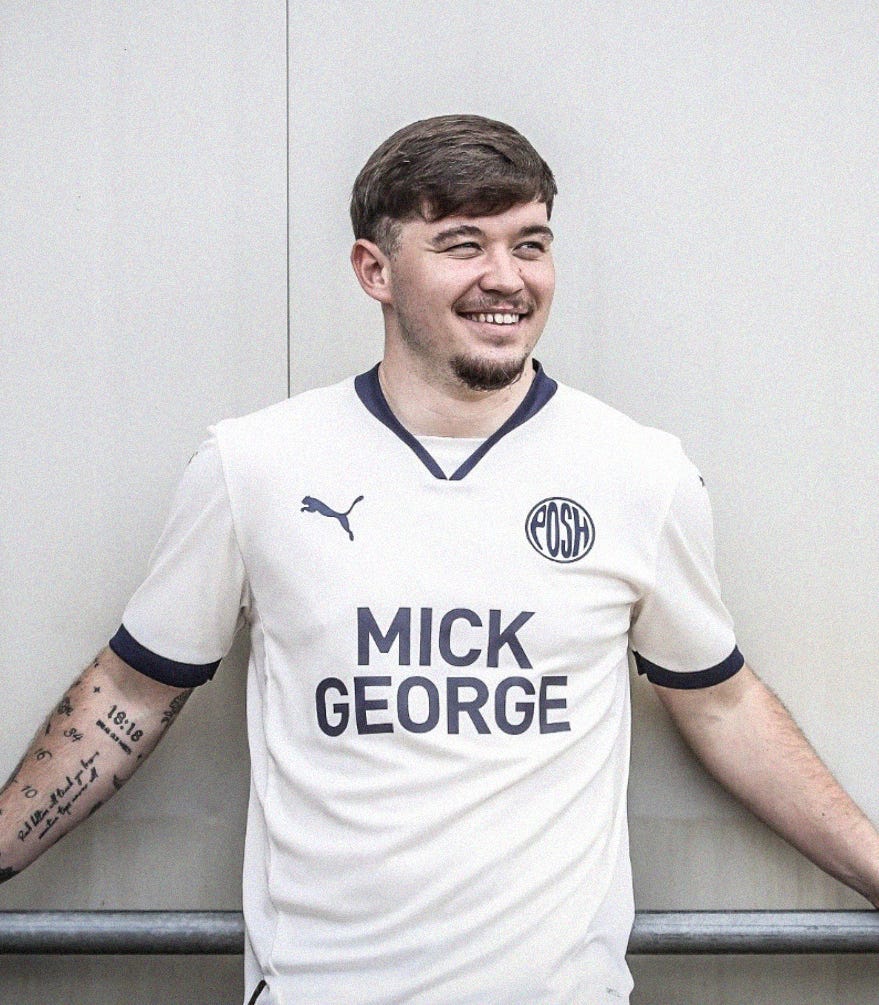 A photo of Chris Conn-Clarke in a retro-inspired white Peterborough United shirt; he's leading against a wall, looking to the left, with a smile on his face