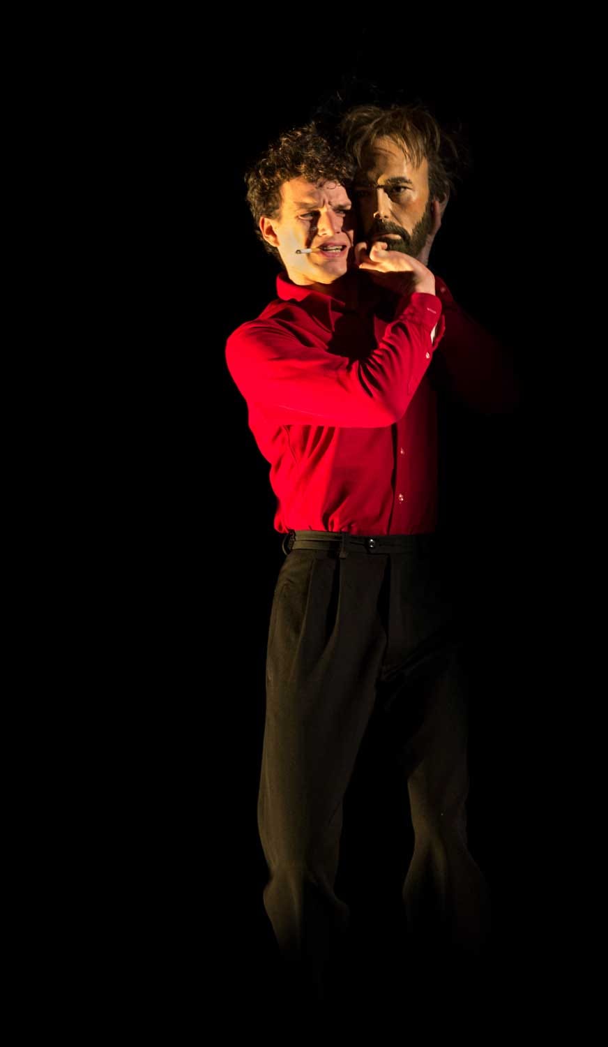 An actor in a red shirt holding a lifelike human head on his shoulder.