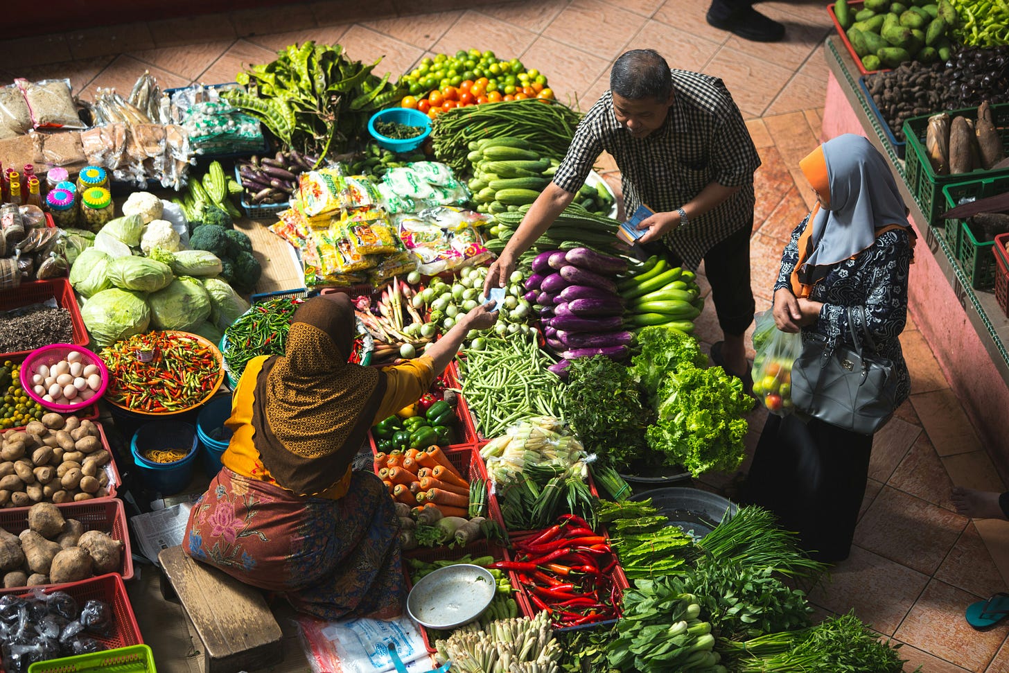 Image: Birds eye view of a woman in a sari and headscarf sitting at her stall surrounded by fresh, colourful vegetables in Siti Khatijah market in Kelantan, Malaysia. A man is reaching out to pay her while his wife holds a bag of produce. © Alex Hudson 2019