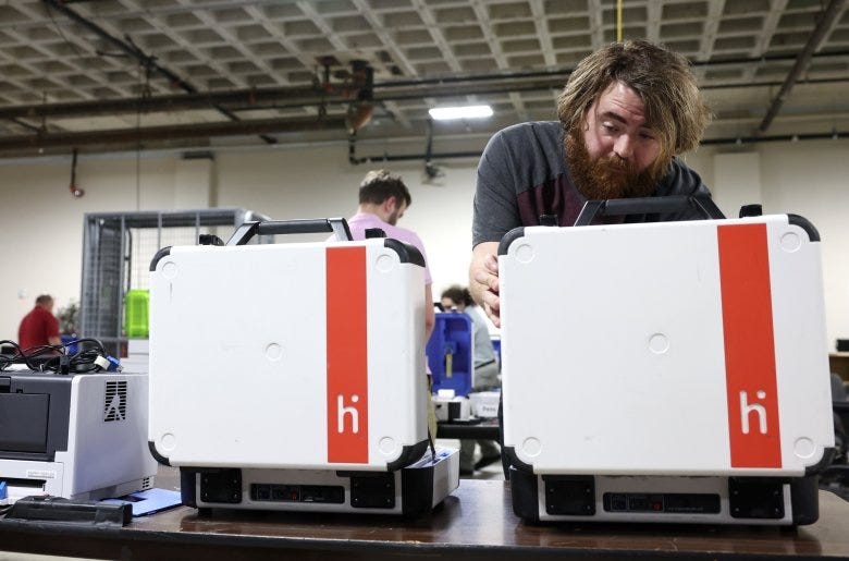 Thomas Castulik is leaning over voting equipment that looks like a small opened suitcase, white with an orange stripe and the company's logo. In the background, other election staff are at work.