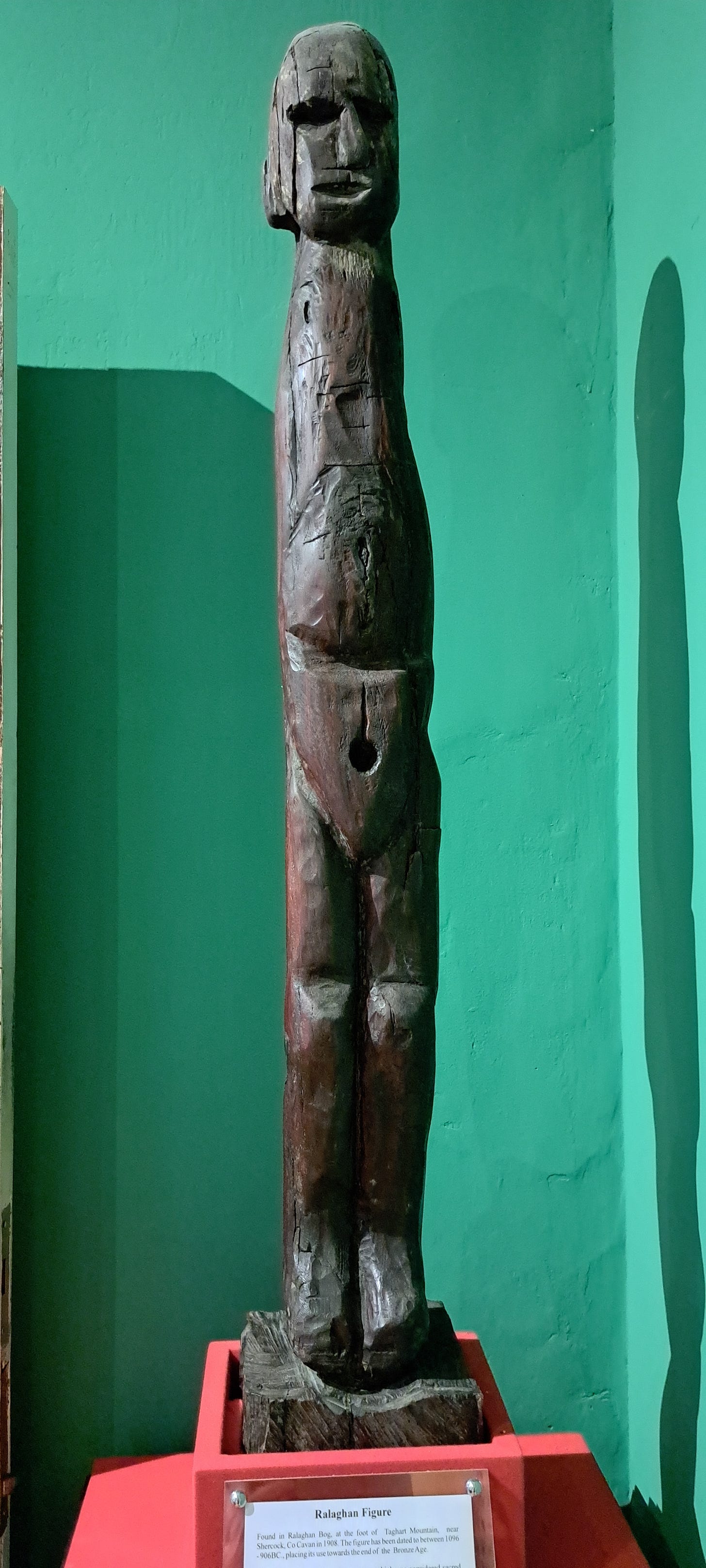 A tall narrow human figure carved from dark wood set on a red plinth and located against a deep green wall. This enigmatic carving is very roughly hewn with only the barest suggestion of arms, but its most significant feature is the round hole in the pelvic area.