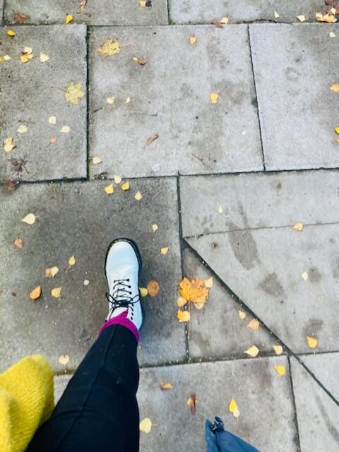 A birdseye view of a grey concrete pavement with a leg shown walking from the bottom left of the image, it has what looks like black leggings a dark hot pink sock and white DM boots. Against the pavement are a scattering of autumn leaves in warm oranges and yellows.
