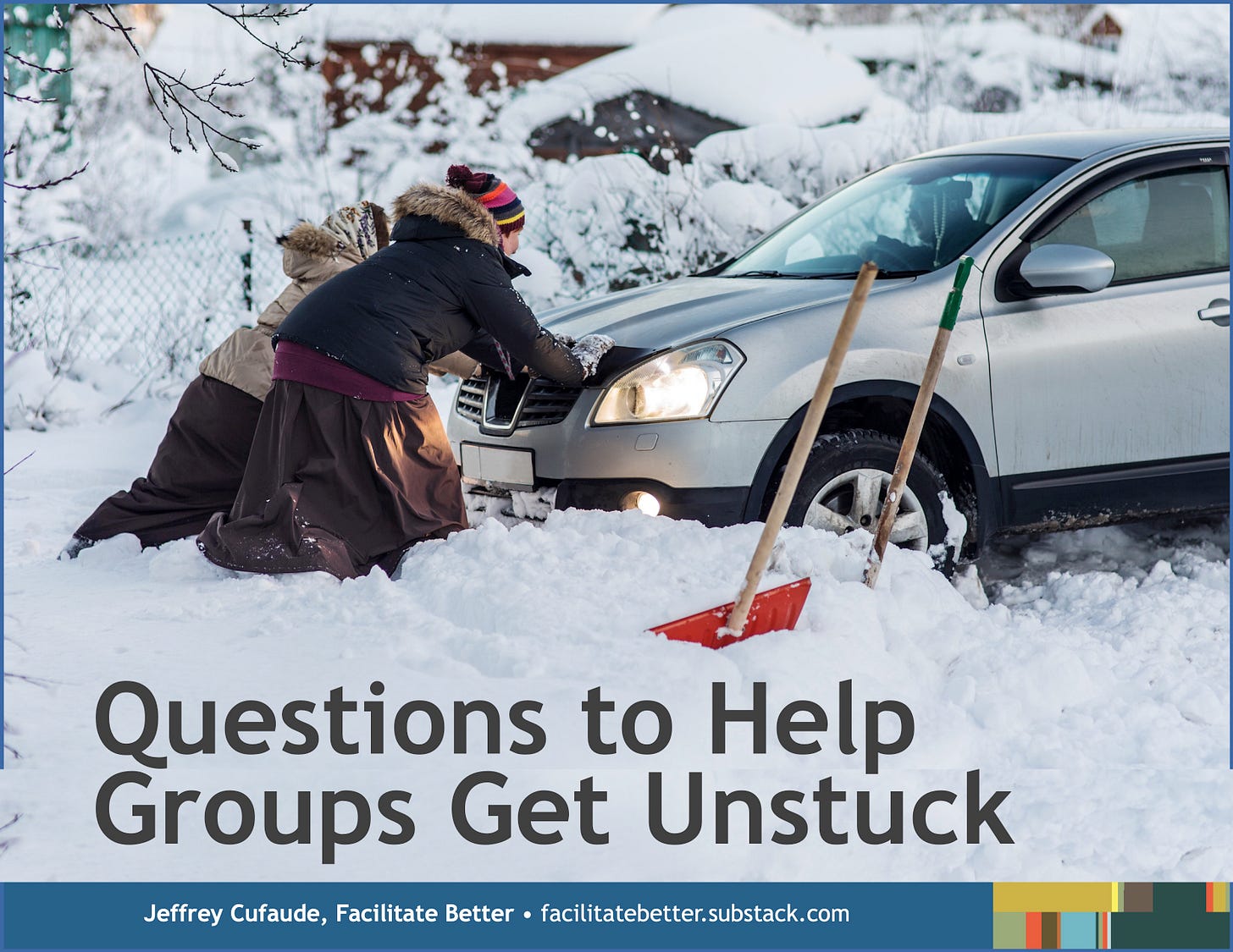 Two people in heavy winter attire push the front of a car stuck in snow. Two shovels stand in the snow beside them.