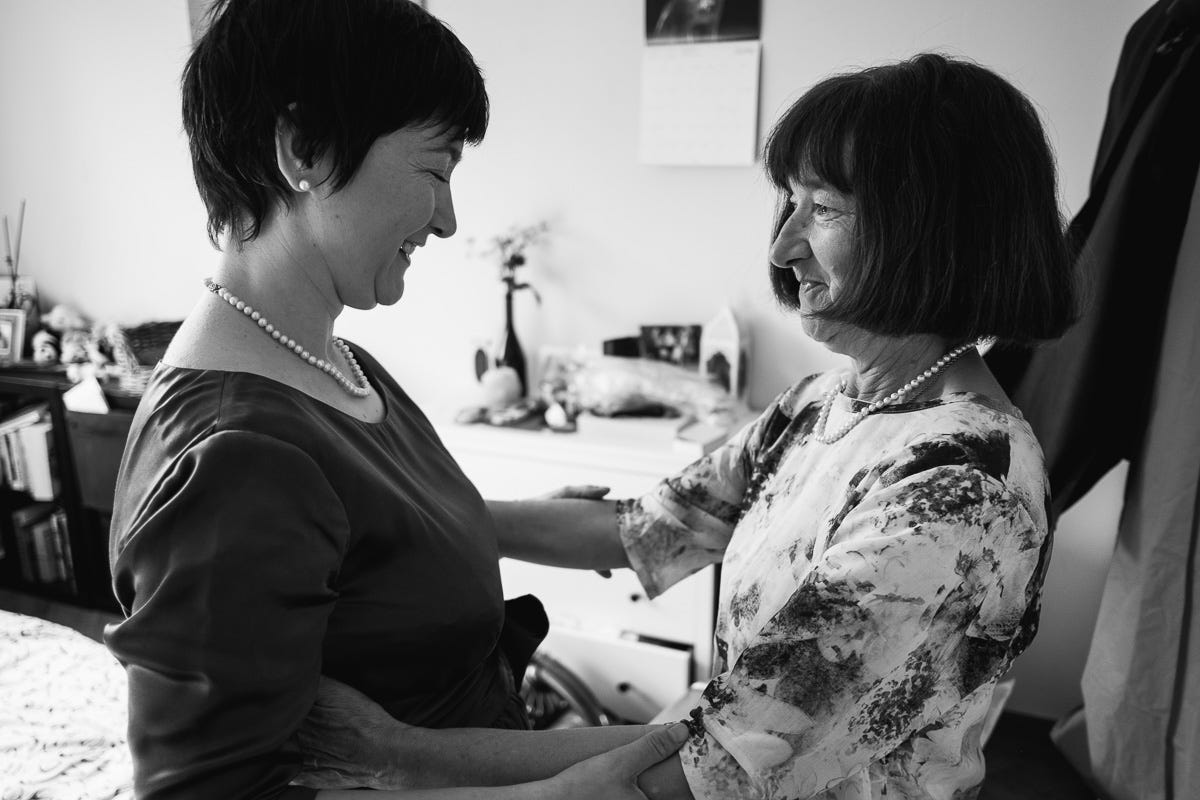 A  black and white image of a mother and daughter embracing on the daughter's wedding day. Mother is about sixty years old, the daughter about forty. The daughter is wearing a dark silky dress and the mother a flower-patterned blouse. They both have pearl necklaces and are smiling