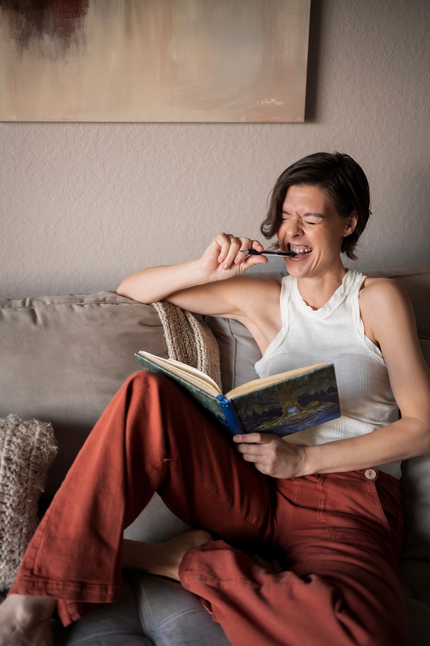 Woman with orange pants and a cream colored tank top sits on a couch with her journal. She is biting on the end of a pencil as if frustrated by writer's block.