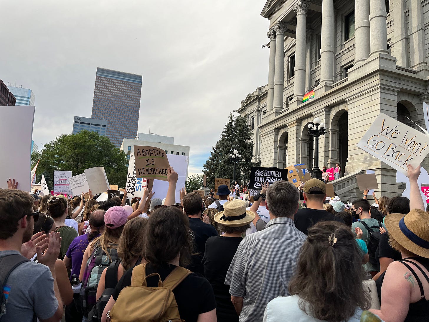 Protestors gather in front of a capitol building. One of them is holding up a sign that says, "Abortion is Healthcare." Another is holding a sign that says, "We won't go back."