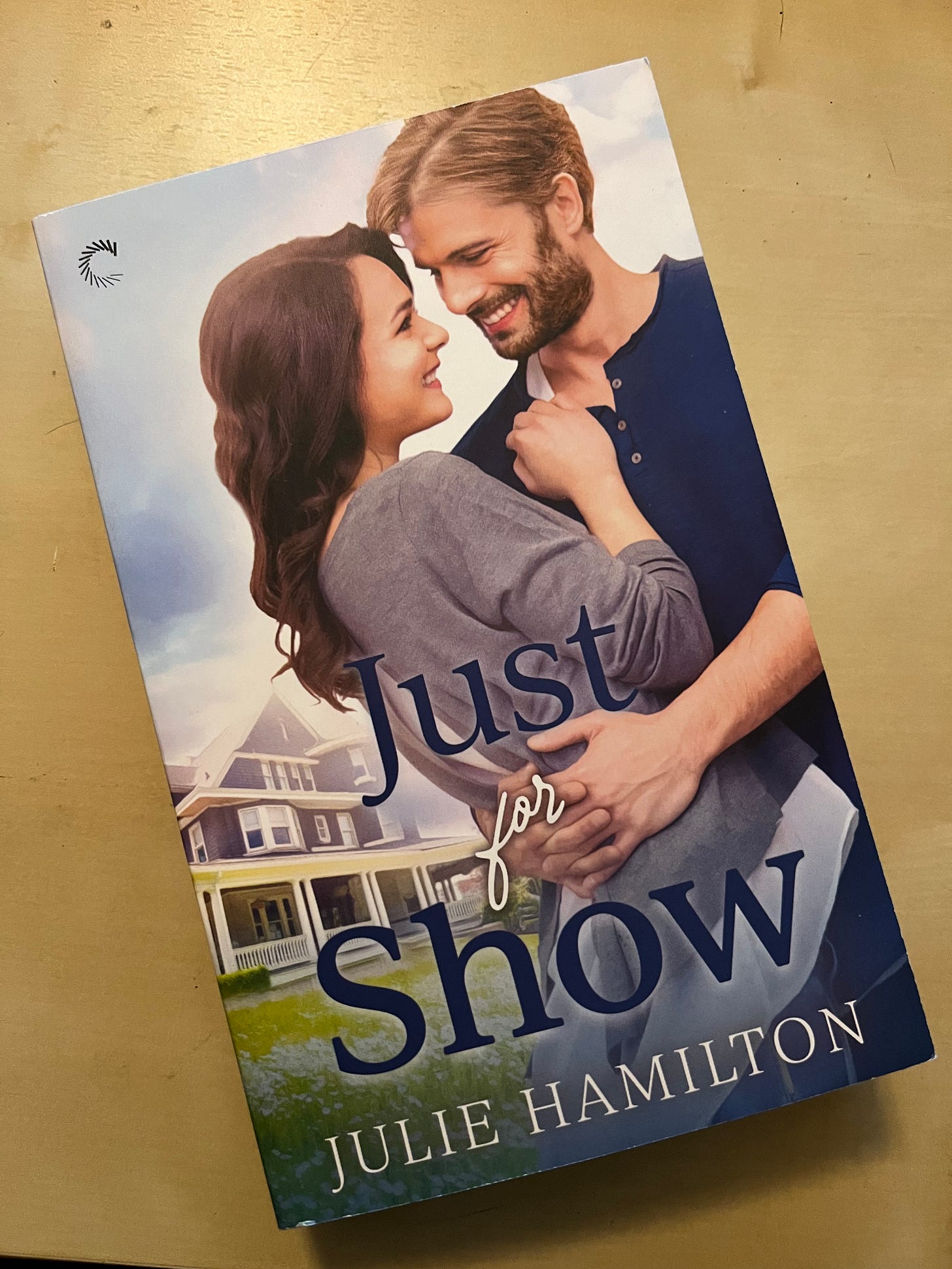 Picture of copy of JUST FOR SHOW by Julie Hamilton on top of a wooden desk, where the cover shows a white couple embracing in front of an old house behind them.