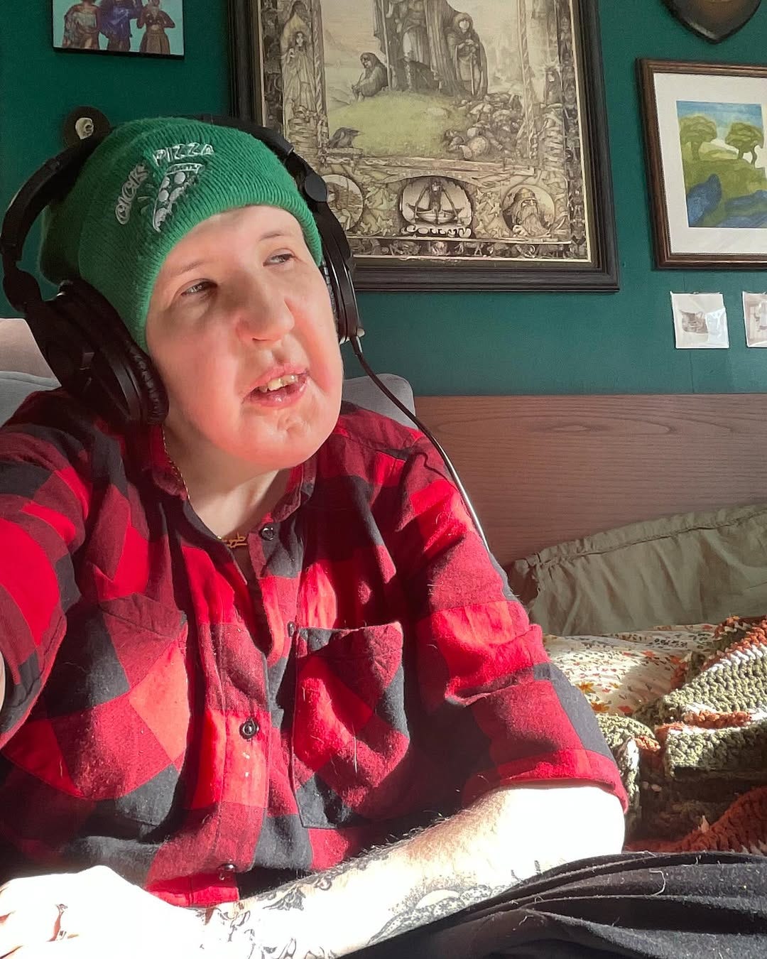 A white queer trans disabled person wearing a green beanie hat, headphones, a red and black plaid button-down shirt. They are sitting on a bed with artworks hanging on the wall behind. They look out to the middle distance in contemplation.