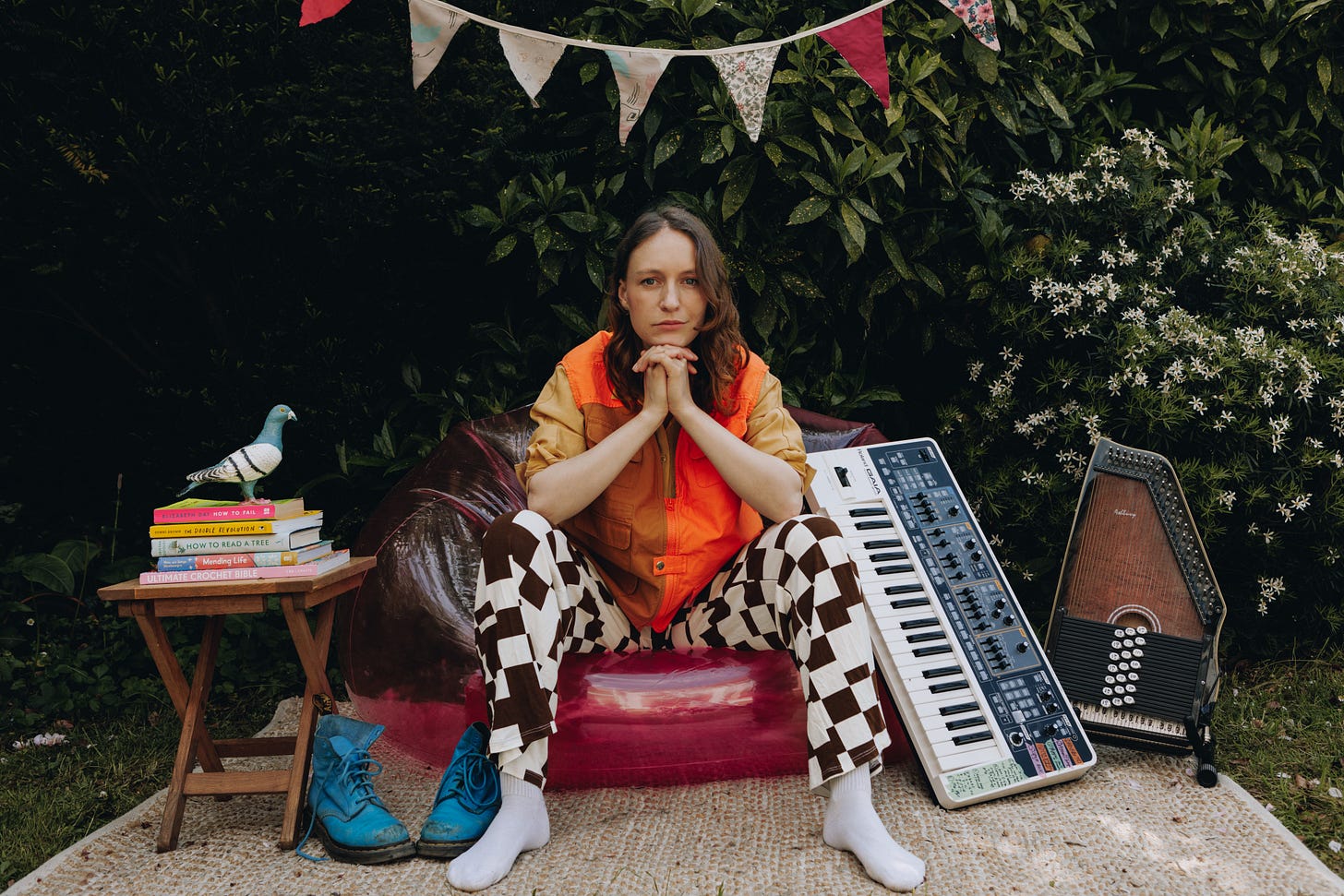 A woman with brown wavy hair sits in a pink inflatable armchair, resting her elbows on her knees and her chin on her folded hands. She is dressed in an orange and brown vest, a yellow shirt, and brown and white flared checkerboard trousers. Behind her, there is lush green foliage and colourful bunting hanging above. To her left, a small wooden table holds a cast iron pigeon atop a stack of books. To her right, there is a white synthesizer and an autoharp.