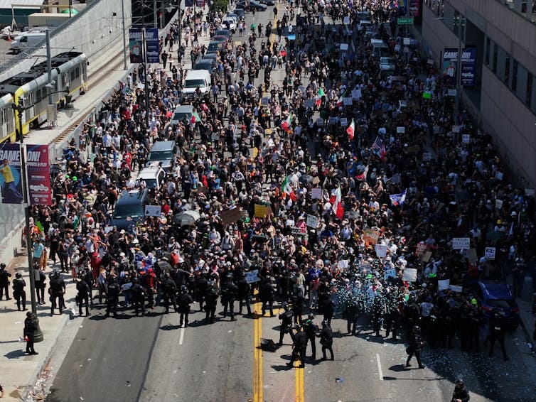 A large crowd of people stand together on a street, with a line of people in black standing close to them. A large crowd of people stand together on a street, with a line of people in black standing close to them.