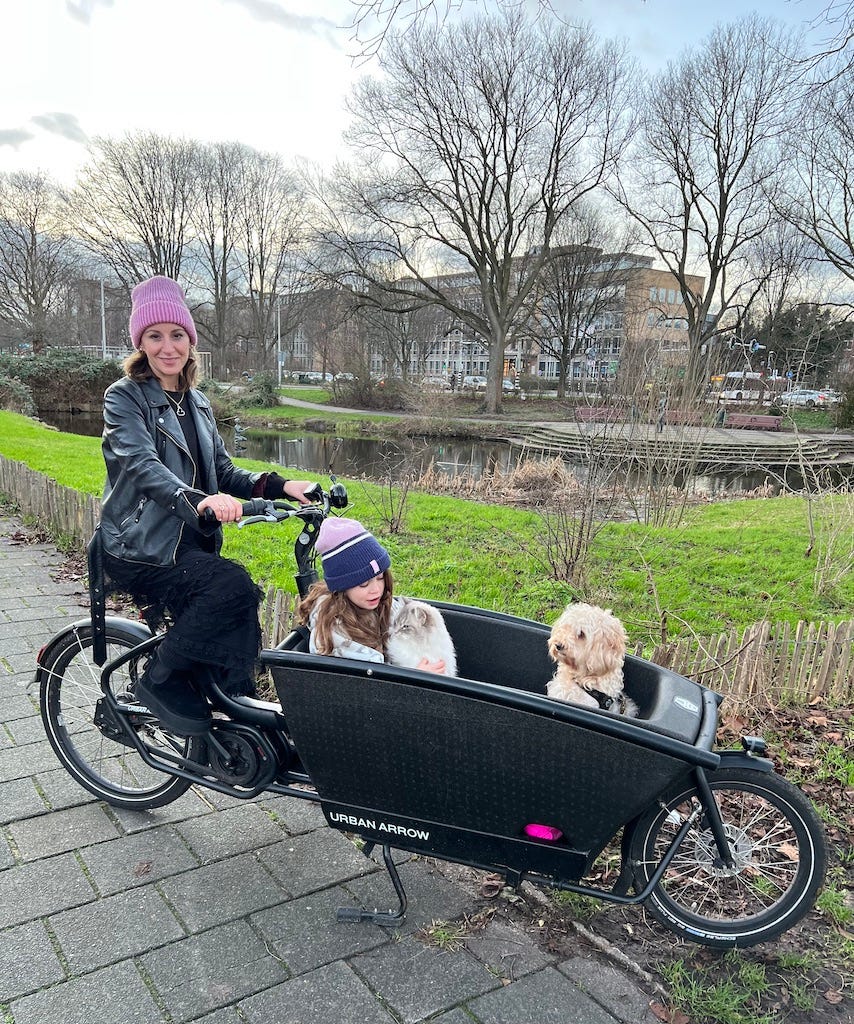 Anna sat on the saddle of a cargobike. Inside the front box of the cargobike are her daughter Zena, their dog and their cat. 