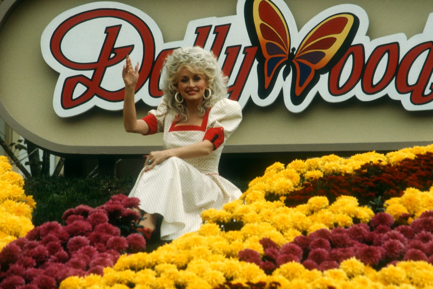 Dolly Parton is sitting among red and yellow flowers waving in front of sign that reads "Dollywood"