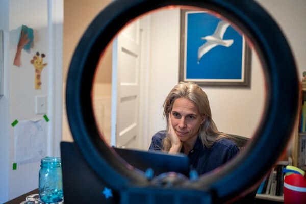 A view through a ring light of Ms. Delano looking at her computer in her office. A picture of a gull hangs on the wall behind her.