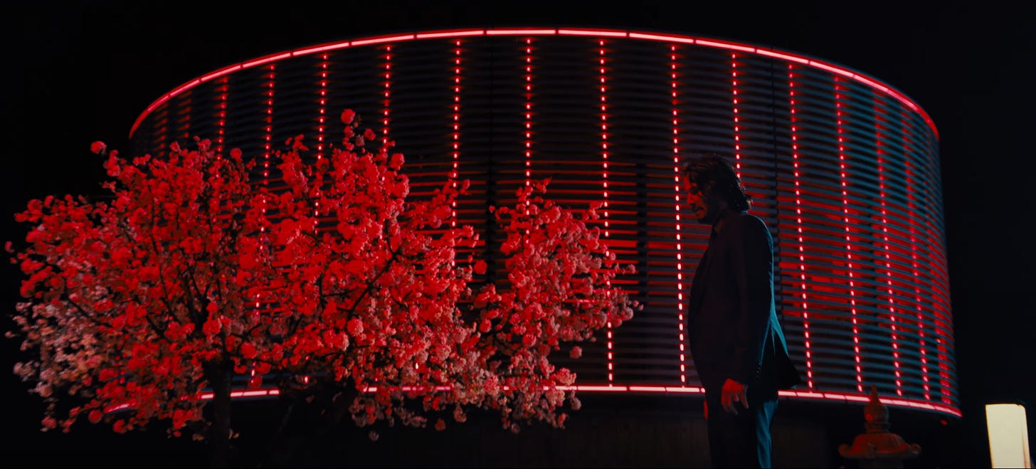 Movie still from John Wick: Chapter 4. John Wick walks outside an Osaka hotel rooftop, a cherry blossom tree in the background drenched in red city lights