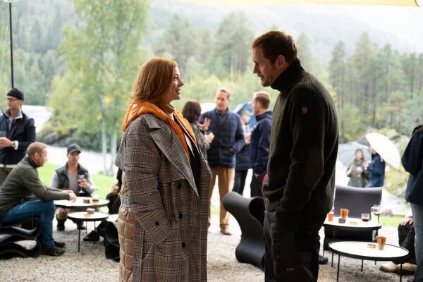 A woman with red hair wearing an oversized gray plaid trench coat speaks to a man wearing a dark colored turtleneck.