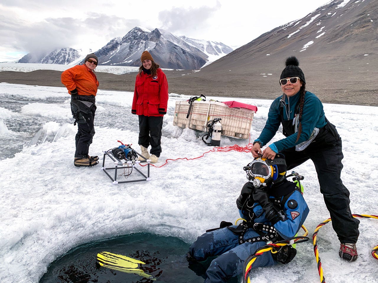 Four researchers in the process of preparing for a dive. One is wearing scuba gear including a full helmet that obscures their face. They are sitting at the edge of a hole in the ice with their feet in the water. The other three wear parkas, hats, and crampons. One is standing above the diver and adjusting something on the dive helmet. They are all looking at the camera and smiling.