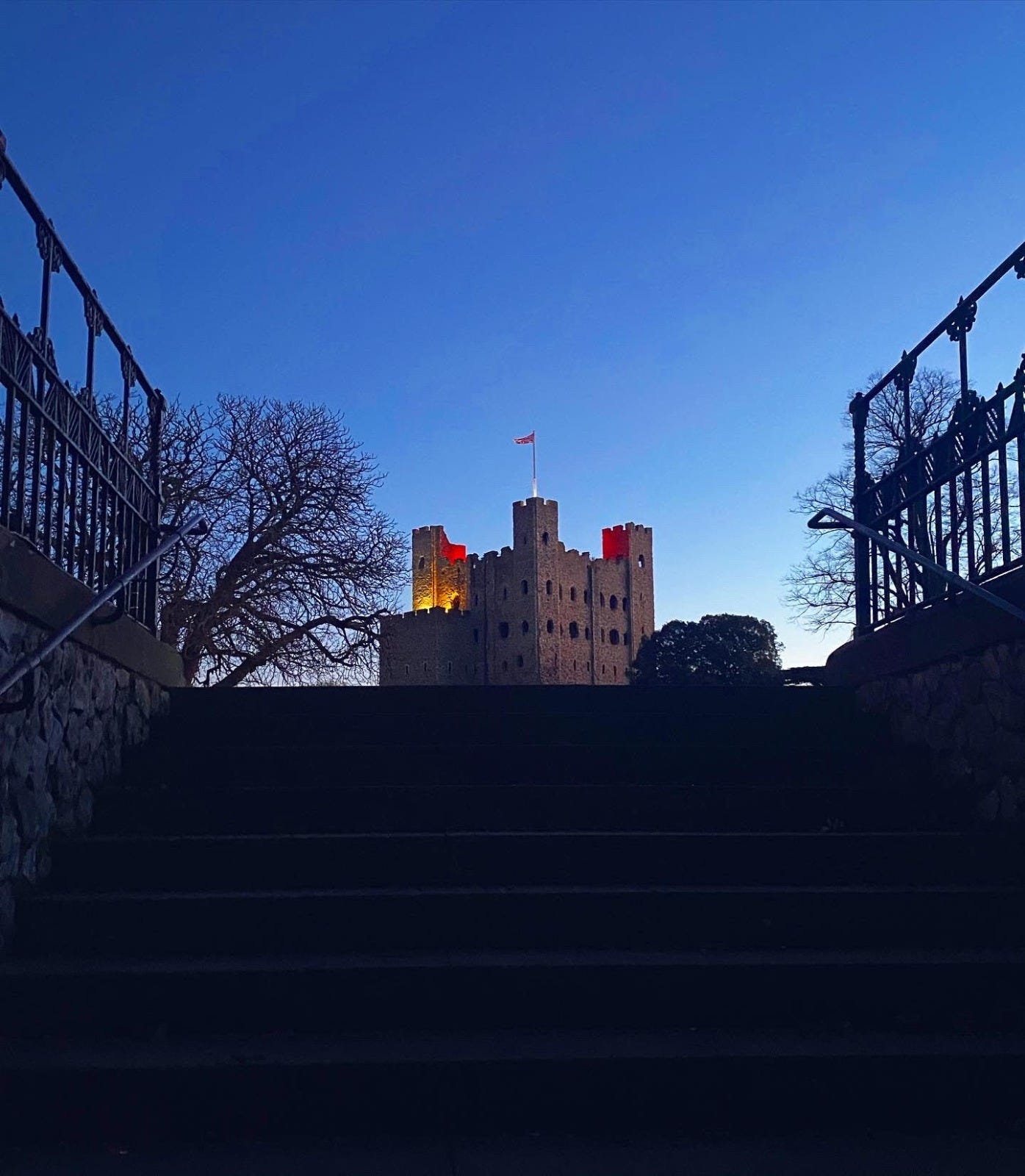 The stairs leading up to Rochexter castle against a picturesque blue sky. It is early evening, when the sky begins to darken. There are trees either side of the castle. The castle itself is built of stone, and lit by a soft red light.
